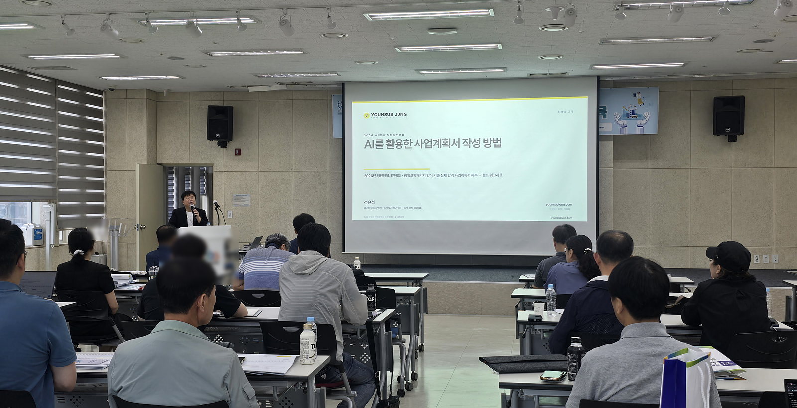 강의실 앞 화면에 「AI를 활용한 사업계획서 작성 방법」 표지 슬라이드가 떠 있고, 수강생들이 앉아 강사를 보고 있다
