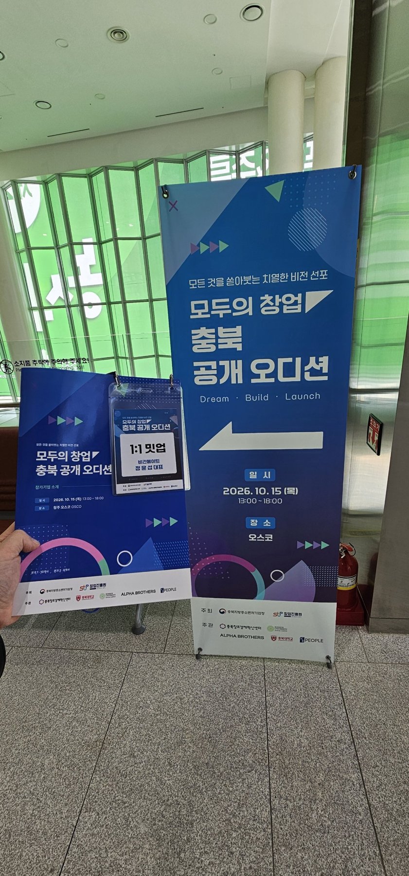 1:1 밋업 명찰을 들고 모두의 창업 충북 공개 오디션 배너 앞에 선 모습
